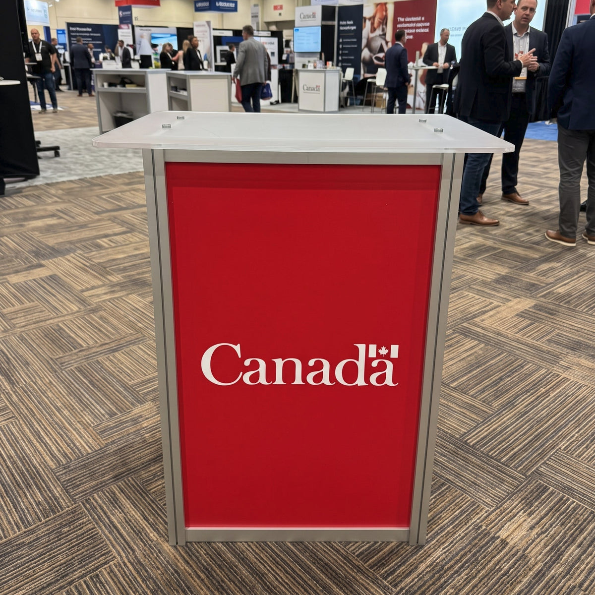 A freestanding rectangular booth counter or display table with a bright red front panel and silver metal frame, printed with the white text 'Canada'. The product is photographed at a trade show or exhibition hall with carpeted floors, other booths, and several people standing in the background.