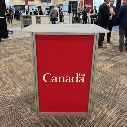 A freestanding rectangular booth counter or display table with a bright red front panel and silver metal frame, printed with the white text 'Canada'. The product is photographed at a trade show or exhibition hall with carpeted floors, other booths, and several people standing in the background.