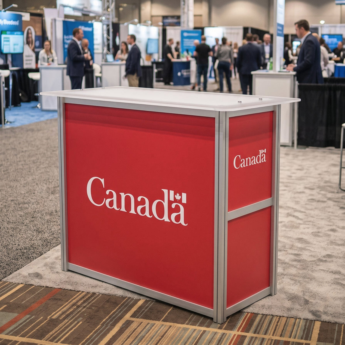 A red and silver Canada booth display table at a trade show or convention, with the word 'Canada' printed in white on the front and side panels. The background shows a busy exhibition hall with booths, people, and overhead truss structures.