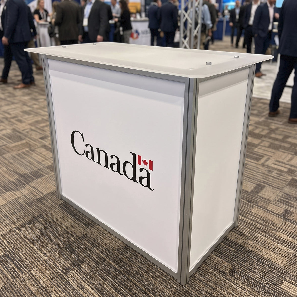 A white and silver trade show booth with the word 'Canada' and a small red maple leaf printed on the front, photographed at a busy convention hall with people and booths in the background.